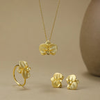 Gold orchid jewelry set featuring a necklace, ring, and stud earrings with textured flower details, displayed on a warm beige background.